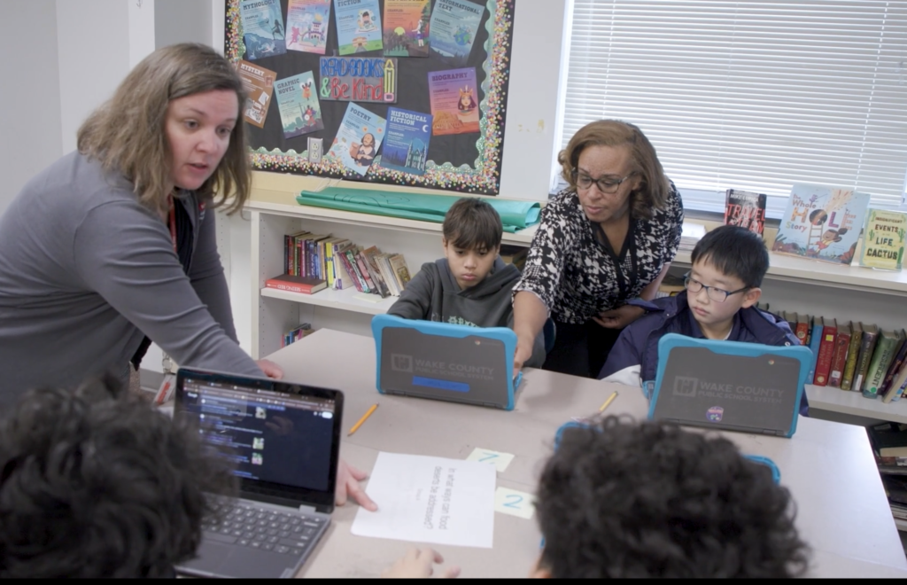 Teachers work with students sitting at a large desk with laptops in front of them.